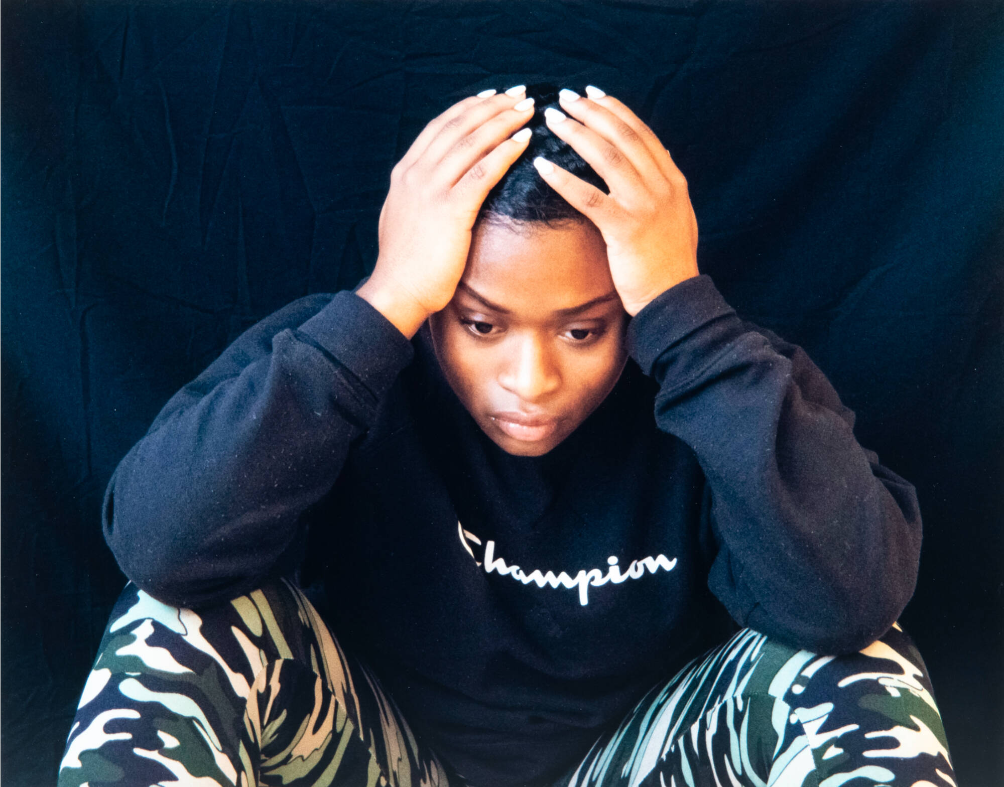 Color photograph of a front view of a person seated on the ground with their elbows on their knees and head in their hands.